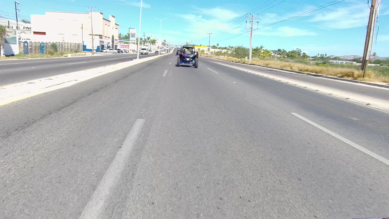 Forward vehicle-level view on a multi-lane road in Los Cabos, Baja California Sur, with cars ahead, roadside buildings on the left, utility poles, bright blue sky, and no visible people.