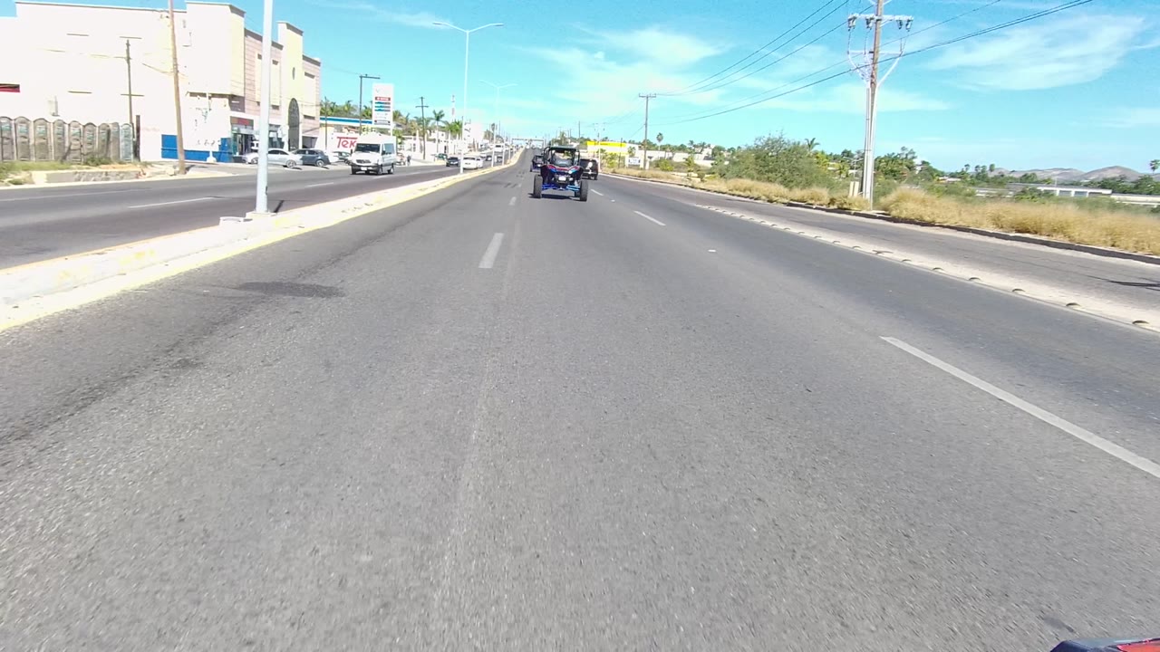 Cars travel ahead on a wide paved road with roadside signs and vegetation.