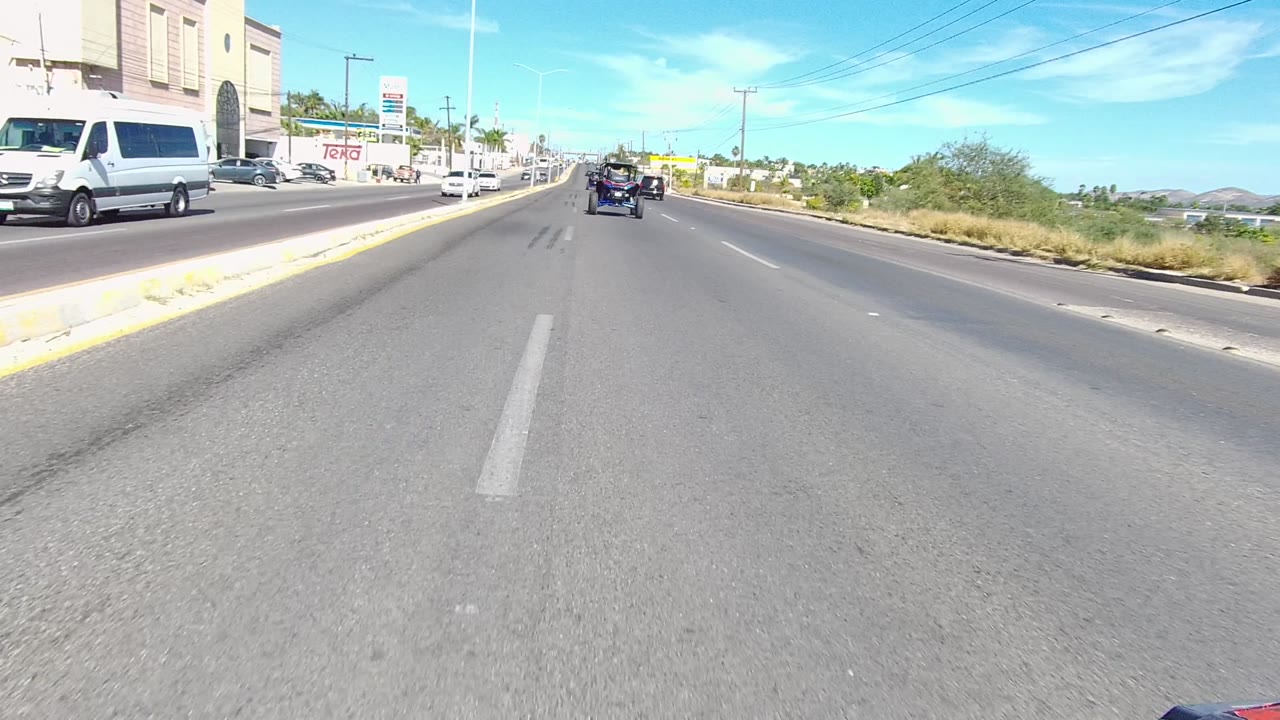 Vehicle-level view along a Los Cabos road with a white van near the left curb, cars ahead in the travel lanes, commercial signage, sidewalk, utility poles, and no visible people.