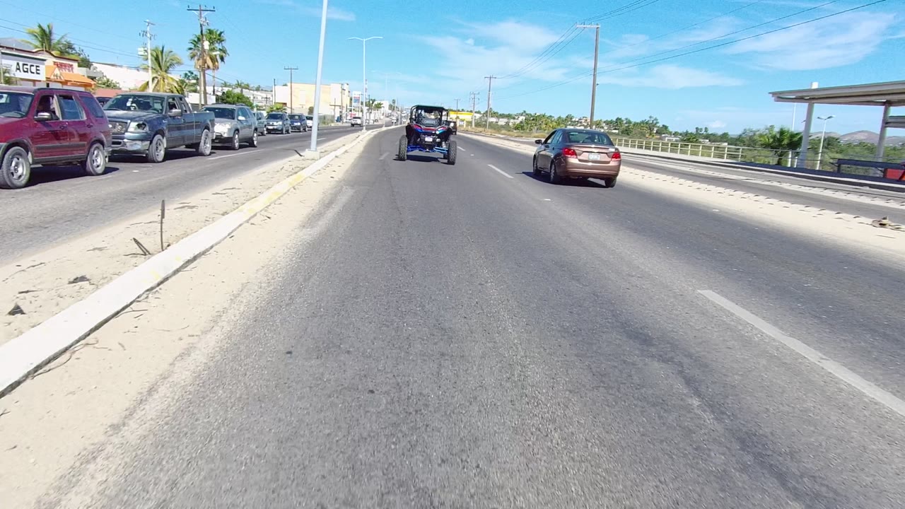 Opening frame of a Los Cabos multi-lane road with cars on both sides, a small dark vehicle ahead, palm trees, low buildings, utility poles, and no visible people.
