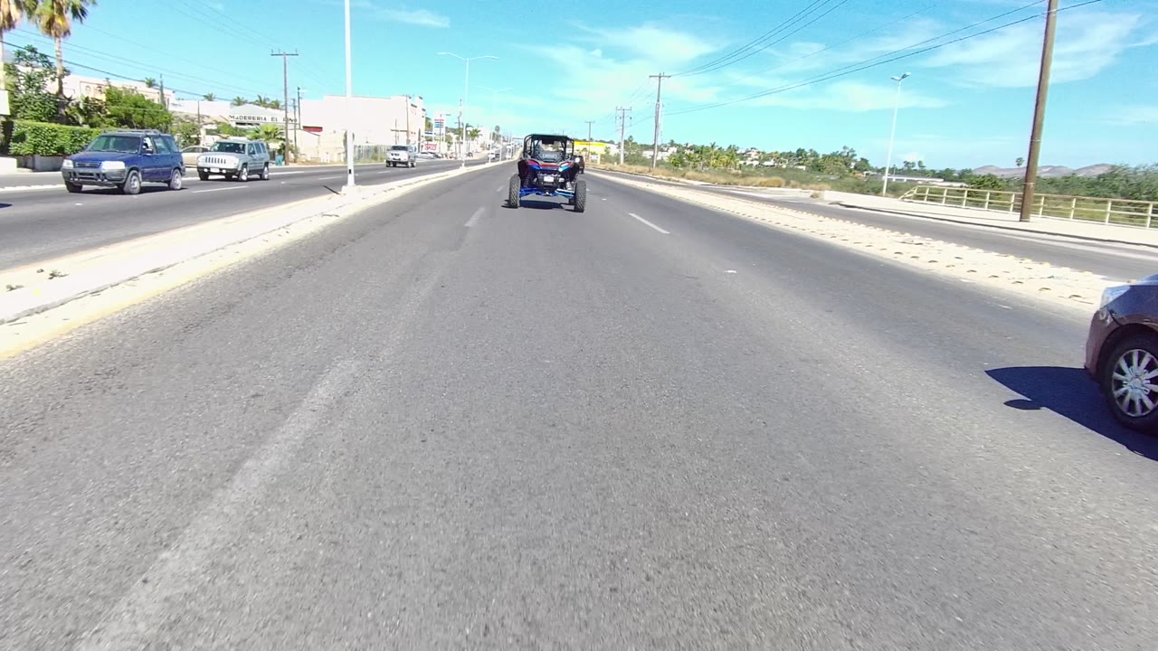 A Los Cabos street view with a brown sedan close on the right, the small dark vehicle farther ahead, roadside buildings, palms, power lines, and no visible people.