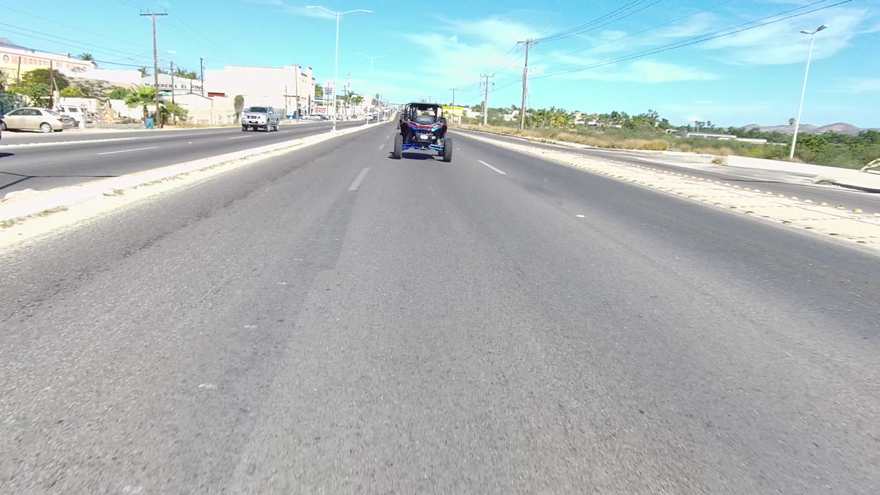 Wide roadway with open shoulder, traffic, buildings, poles, and palms.