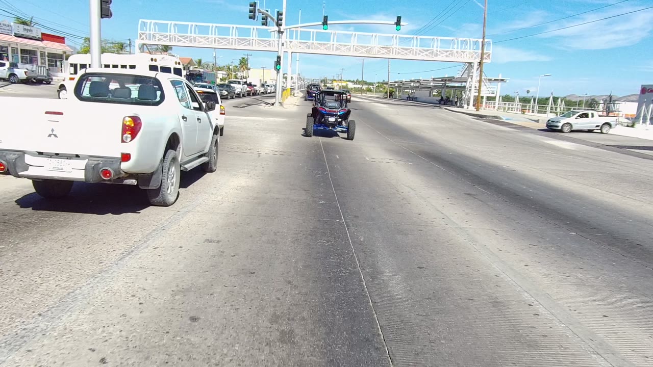 Los Cabos roadway view with a white pickup at left, an off-road vehicle ahead, traffic lights overhead, a white pedestrian bridge, and no visible people.