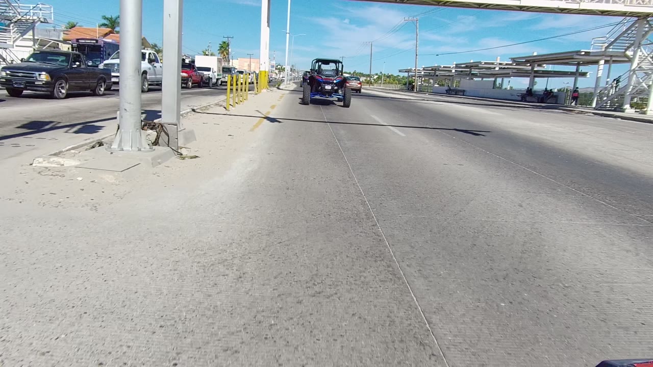 Forward road view in Los Cabos with an off-road vehicle centered ahead, opposing traffic at left, bridge supports at right, bright pavement, and no visible people.