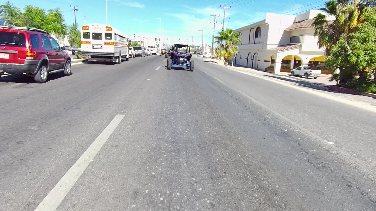 Los Cabos road view with a small open-top vehicle ahead, a bus and red SUV to the left, palms and pale buildings along the street, and no visible people.