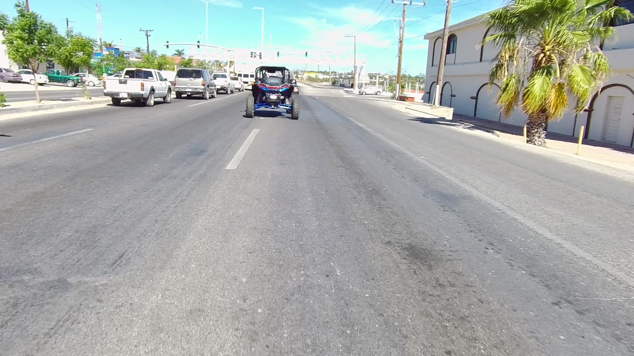 The small open-top vehicle continues ahead toward intersection lights on a sunny Los Cabos road, with trucks, cars, palms, and no visible people.