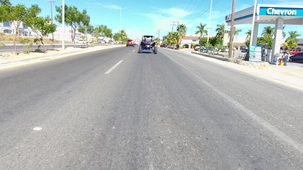 Opening frame of a Los Cabos urban road with a Chevron station sign on the right, vehicles ahead, palm trees, bright blue sky, and no visible people.