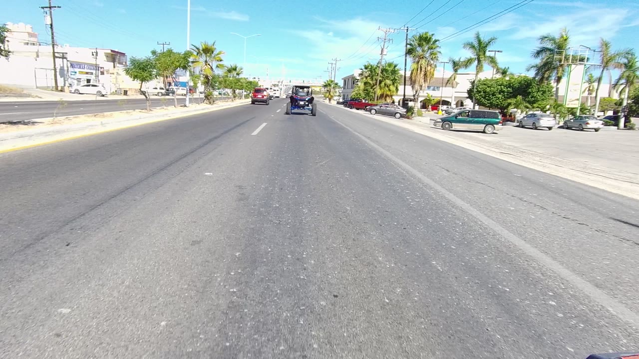 Mid-clip Los Cabos street frame with a dark vehicle centered ahead, palms and commercial-looking buildings along the roadside, parked cars, and no visible people.