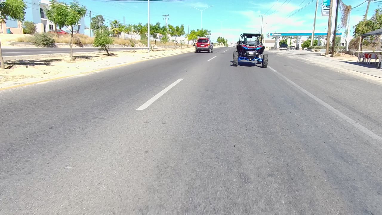 A blue dune buggy drives in the right lane of a wide paved road in Los Cabos, with a red car ahead, roadside trees, buildings, and no visible people.