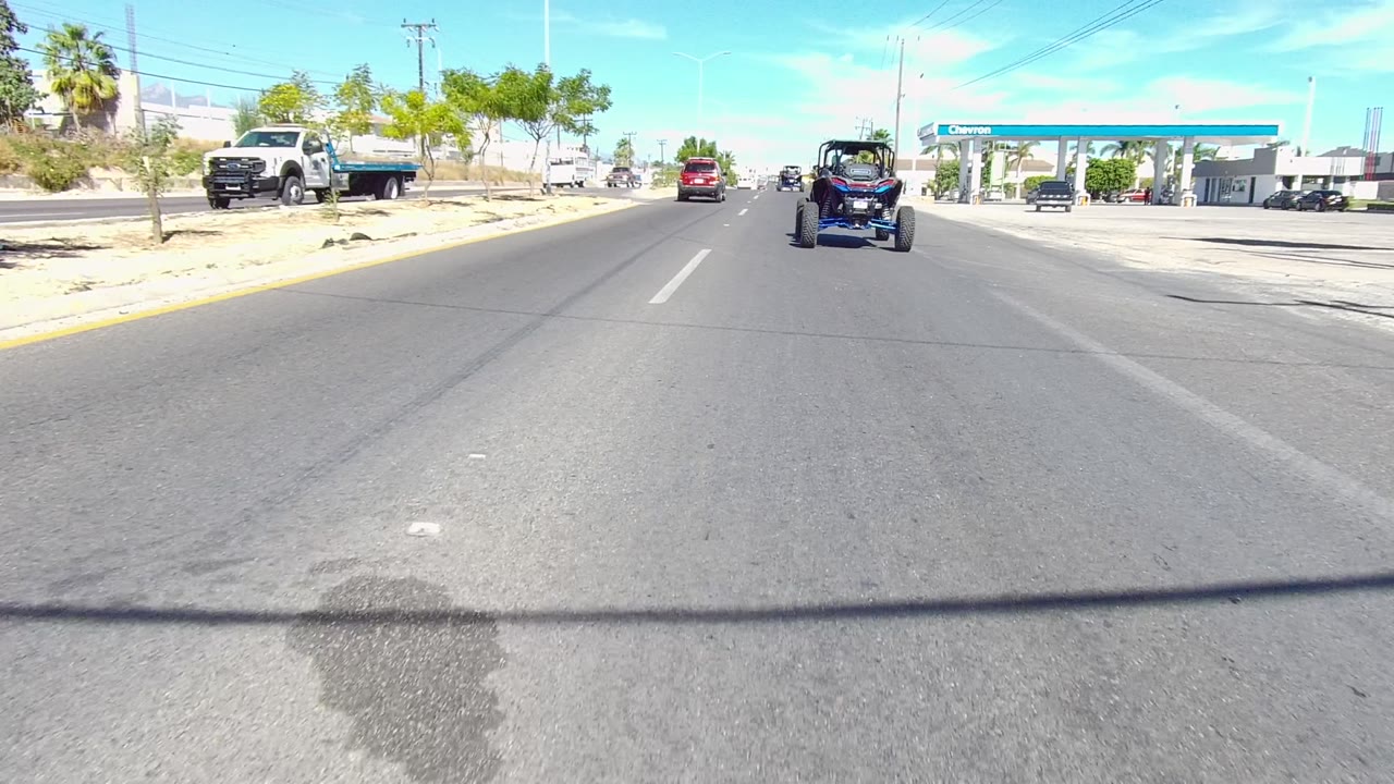 A blue dune buggy follows traffic on a broad Los Cabos road, with the Chevron canopy and parked vehicles visible on the right and no visible people.