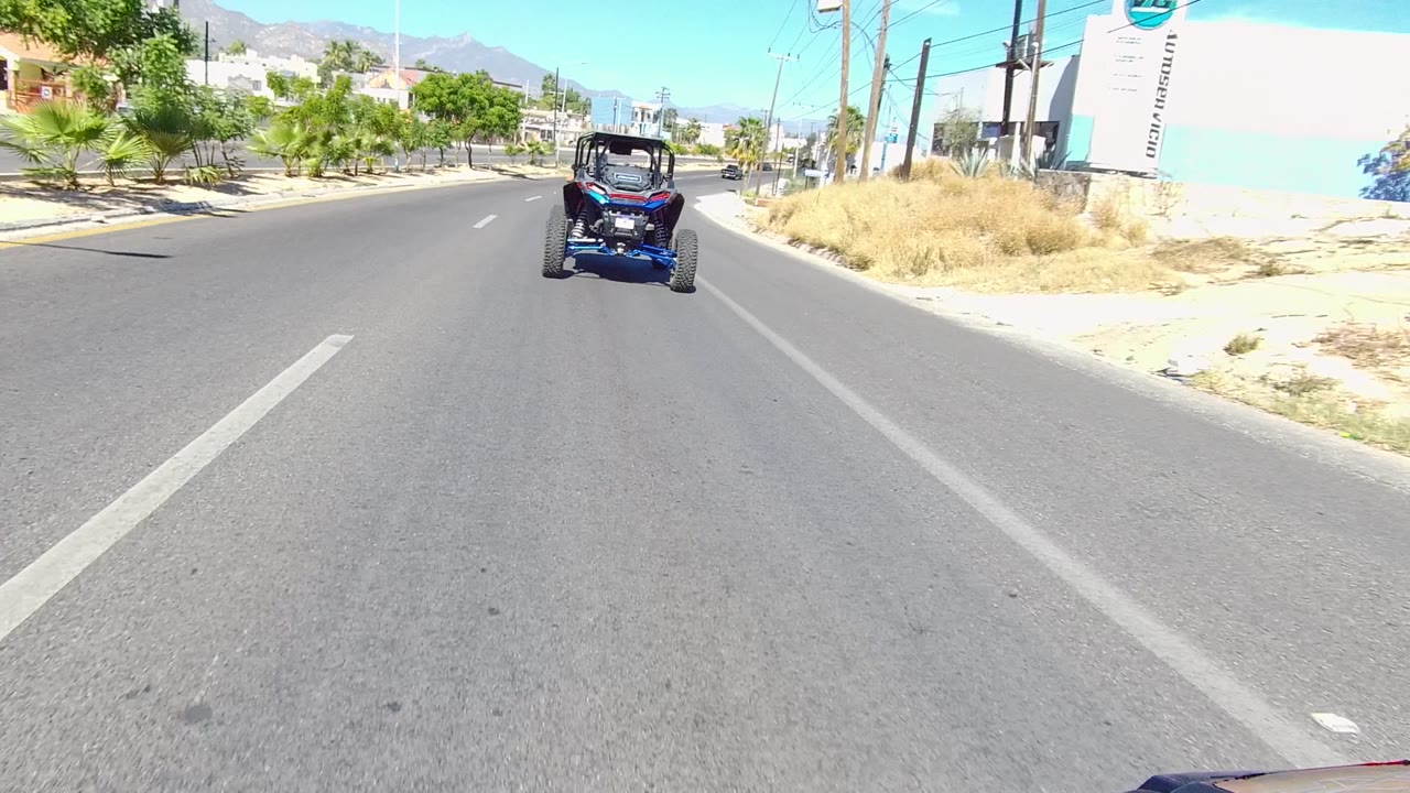 Blue dune buggy drives ahead on a paved road in Los Cabos, Baja California Sur, with dry roadside grass, low buildings, mountains, clear sky, and no visible people.