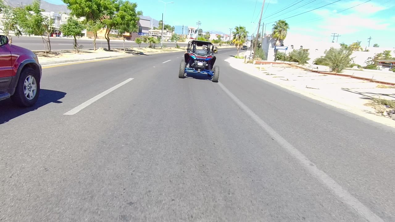 A red SUV appears at the left edge of the roadway as the blue dune buggy remains ahead in Los Cabos, with dry vegetation, low buildings, bright sun, and no visible people.