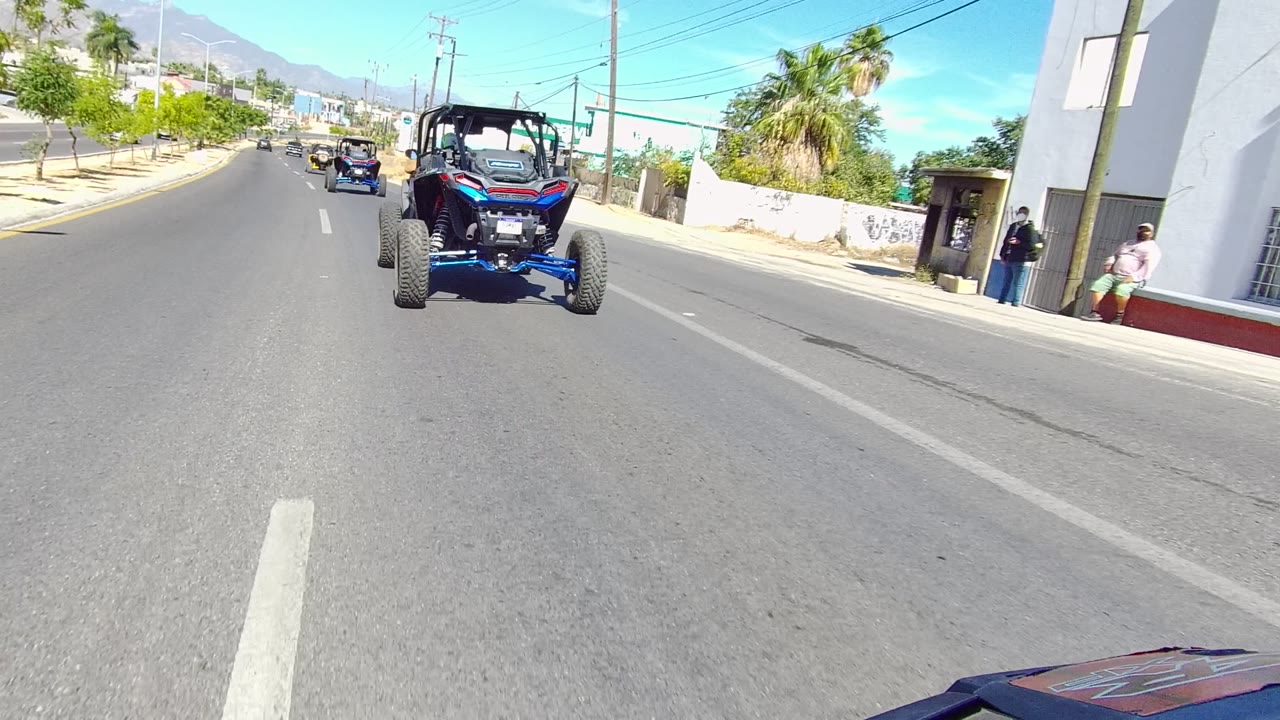 At 0:00 in Los Cabos, a blue dune buggy drives ahead on a paved road, with a white roadside structure, a few visible people, palm trees, and distant mountains in bright daylight.
