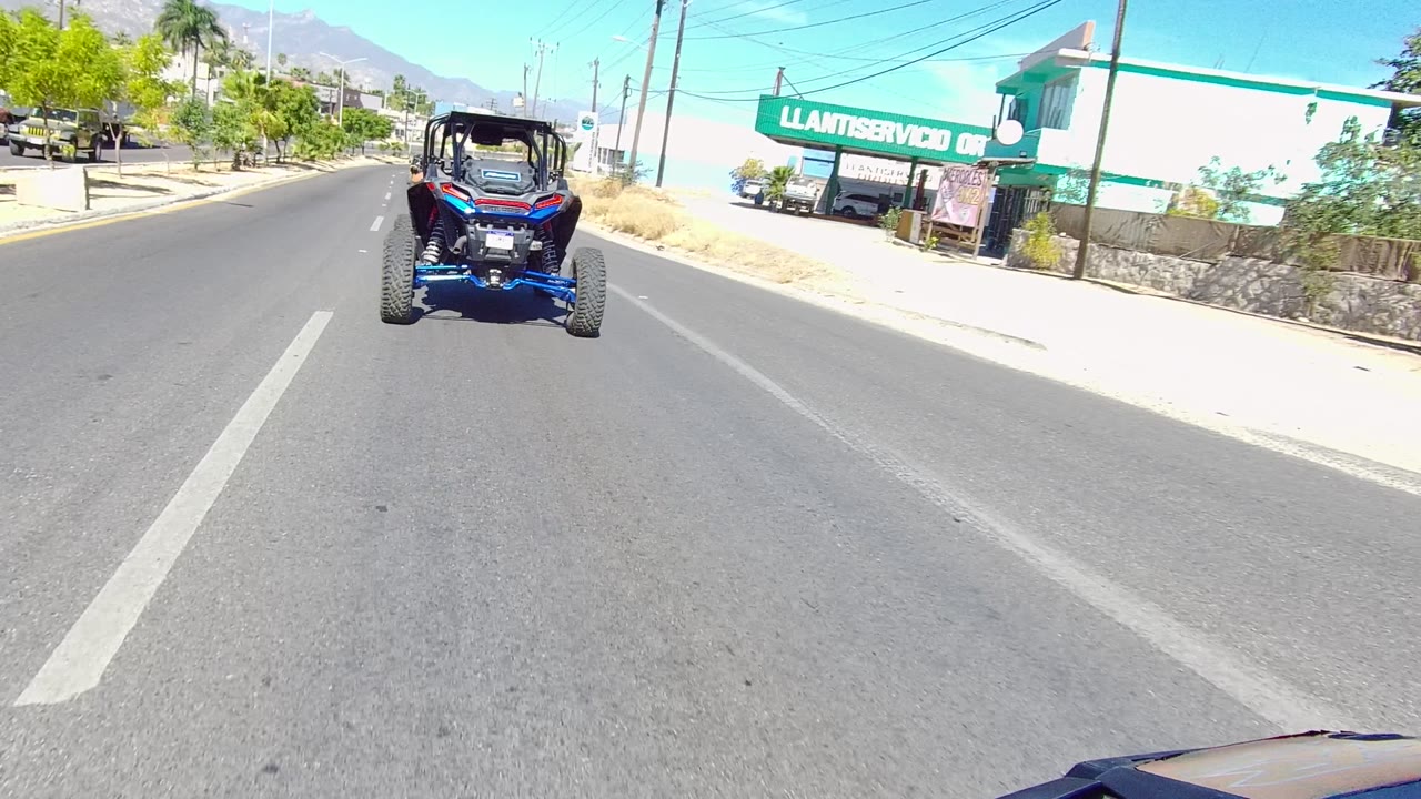 At 0:02 in Los Cabos, the buggy travels away near the center of the lane as a large green tire-service sign, dry grass, utility lines, palm trees, and mountains fill the roadside background.