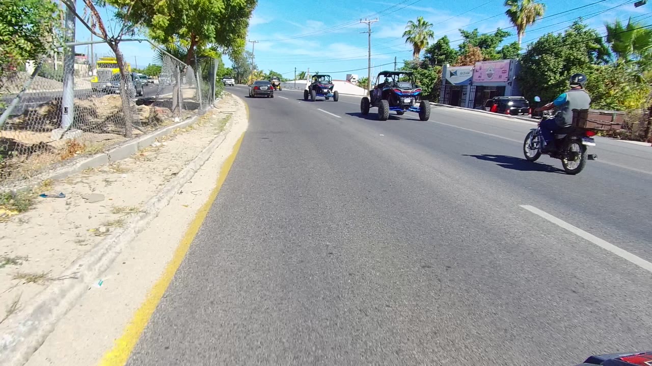 Los Cabos road view with off-road buggies ahead, a motorcycle rider near the right lane, roadside fencing, trees, buildings, and no clearly visible faces.