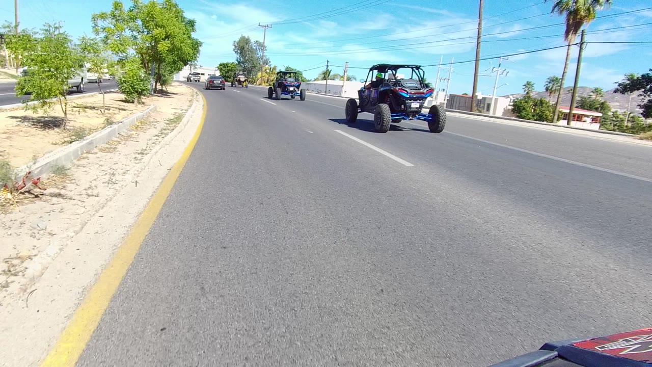 Los Cabos road scene with a rear view of an off-road buggy at right, another vehicle farther ahead, a pale roadside wall, trees, and clear blue sky.
