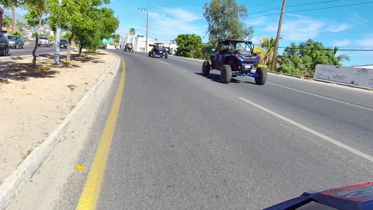 Off-road UTVs drive along a paved road in Los Cabos, Baja California Sur, with trees on the left, white buildings on the right, and no clearly visible people.