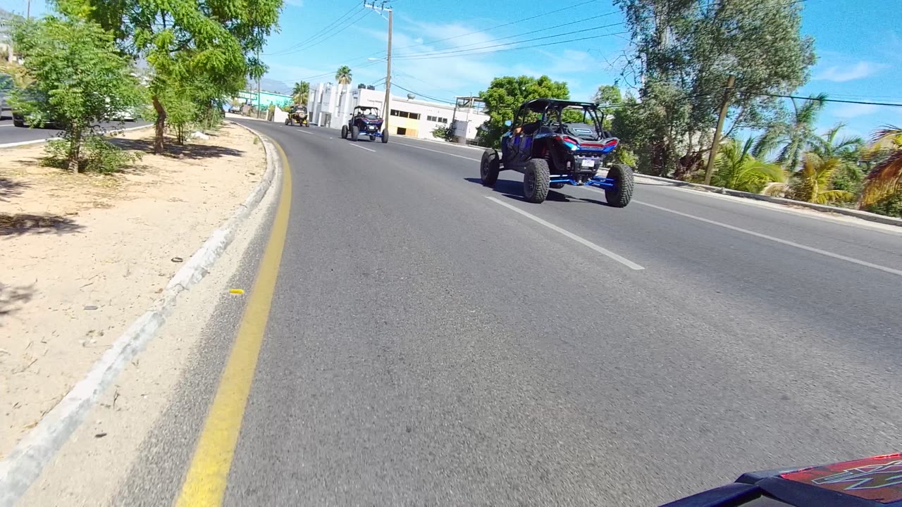 Two off-road UTVs ahead on a curved paved street with trees and low buildings.