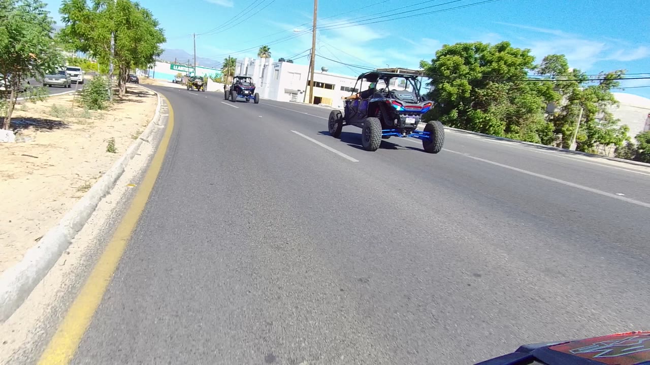 A blue off-road UTV moves farther right in the frame on a Los Cabos paved street, with roadside greenery, white commercial buildings, mountains faintly beyond, and no clearly visible people.