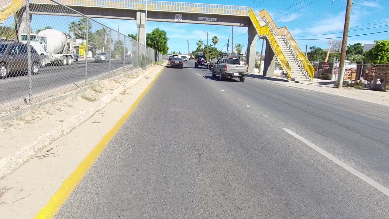Los Cabos city road approaching a yellow-staired pedestrian overpass, with a chain-link fence on the left, cars and a cement truck ahead, and no visible people close to camera.