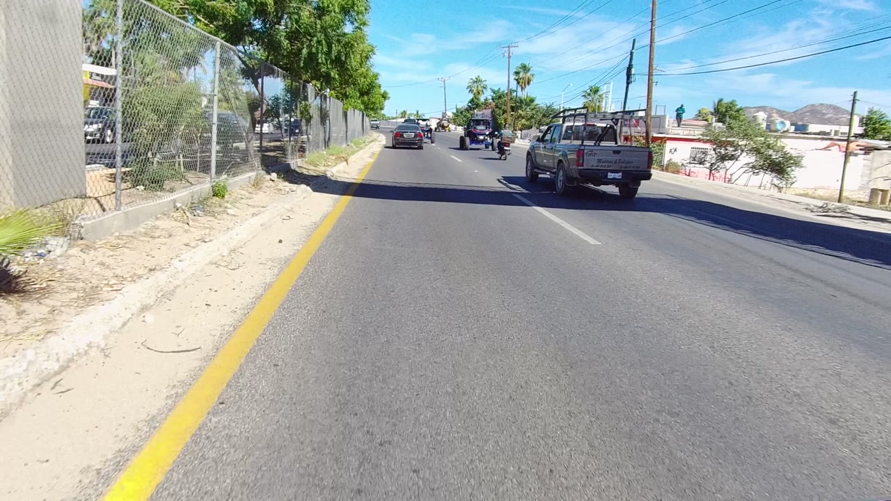 Los Cabos street scene after the overpass, with a pickup truck on the right, cars and motorcycles ahead, chain-link fencing to the left, and no recognizable people.