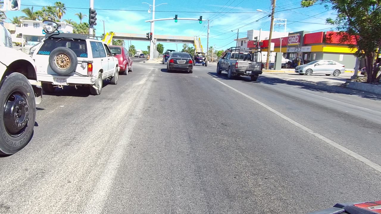 A wide road-level view in Los Cabos, Baja California Sur shows cars and SUVs at a sunny intersection with green traffic lights, storefronts, palm trees, and no visible people.