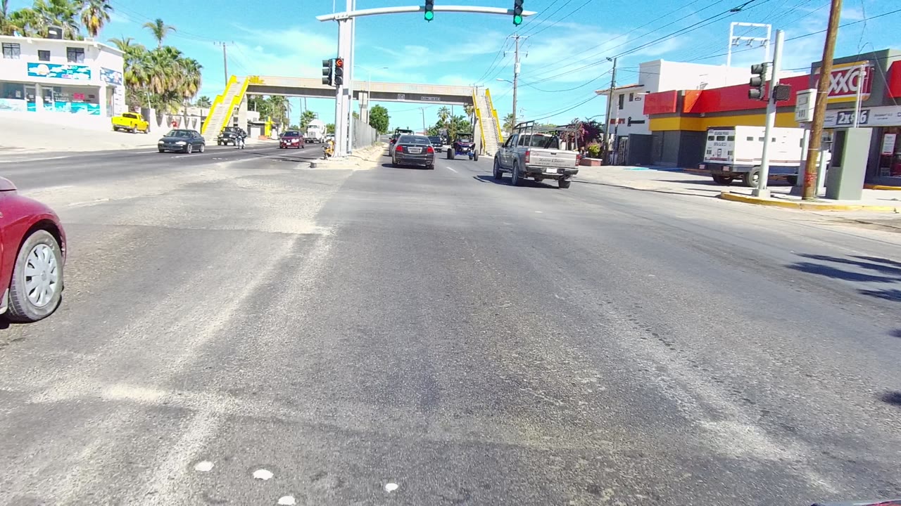 A wide Los Cabos intersection view shows open asphalt lanes, a white car crossing, a pickup truck ahead, a corner storefront with red and yellow signage, and no visible people.