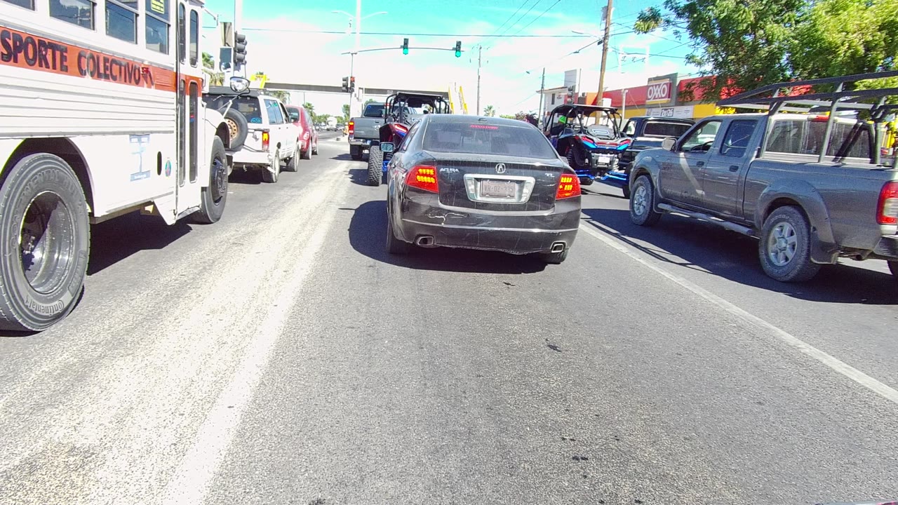 Street-level view of Los Cabos traffic with a bus at the left edge, a dark sedan ahead, a gray pickup to the right, green traffic lights, and no visible people.
