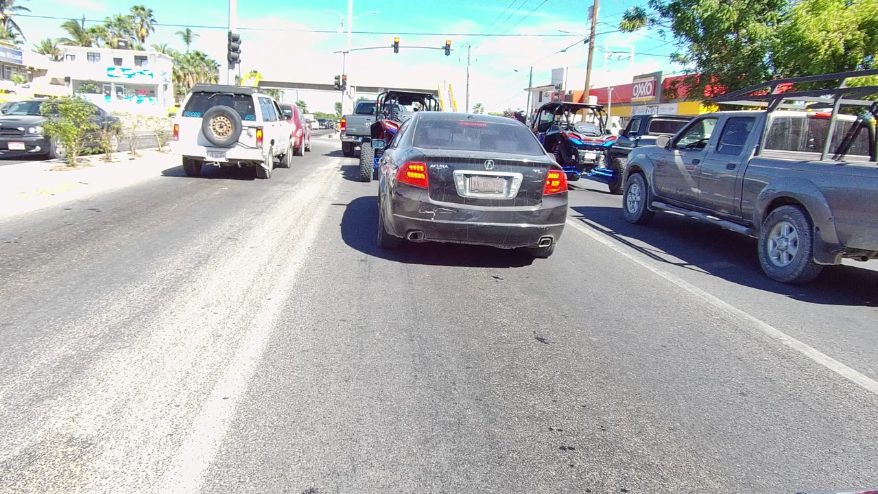 The same Los Cabos traffic lane remains stopped, with the black sedan close to camera, the gray pickup on the right, a red vehicle farther right, traffic signals overhead, and no visible people.