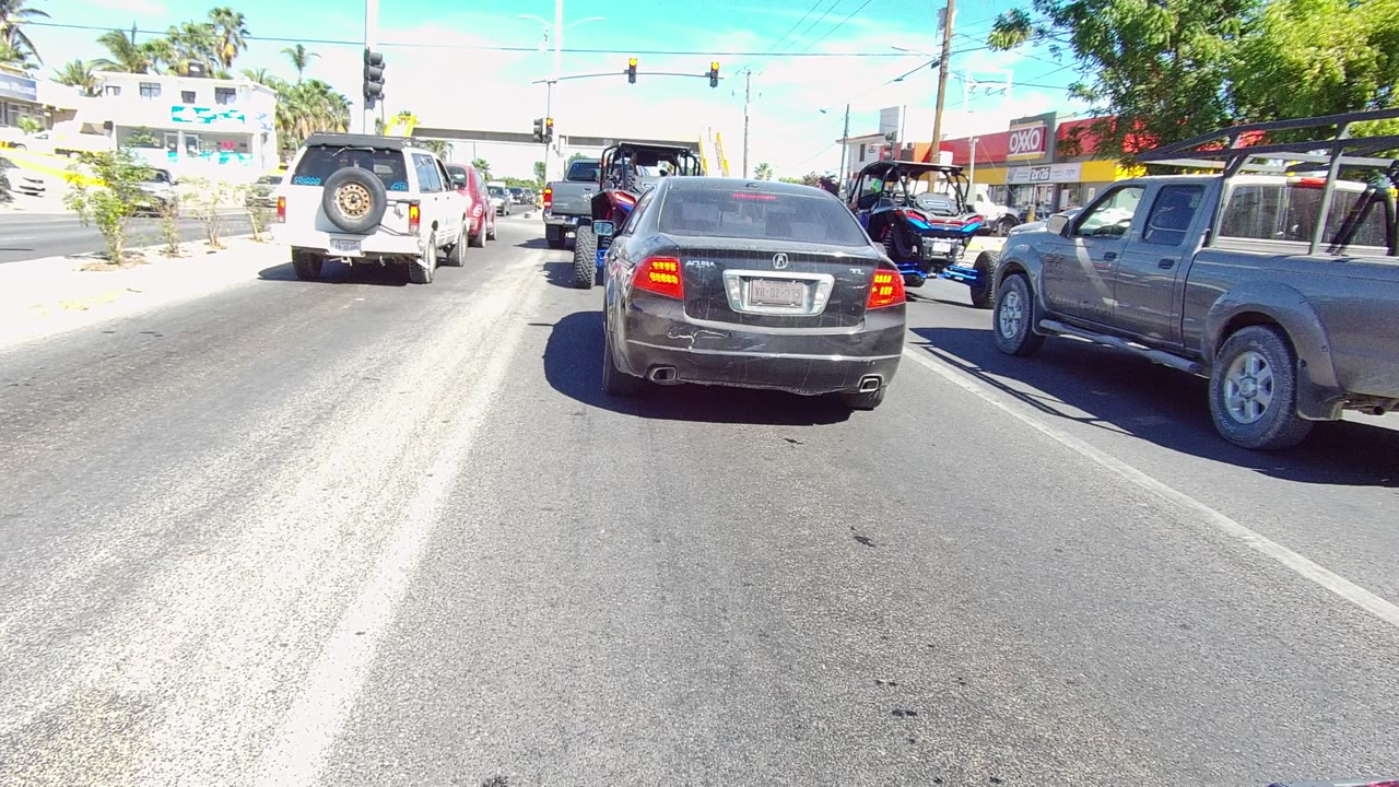 Second frame shows the same Los Cabos roadway with the black sedan near center, pickup trucks to the right, a white SUV in the left lane, overhead signals, bright pavement, and no visible people.