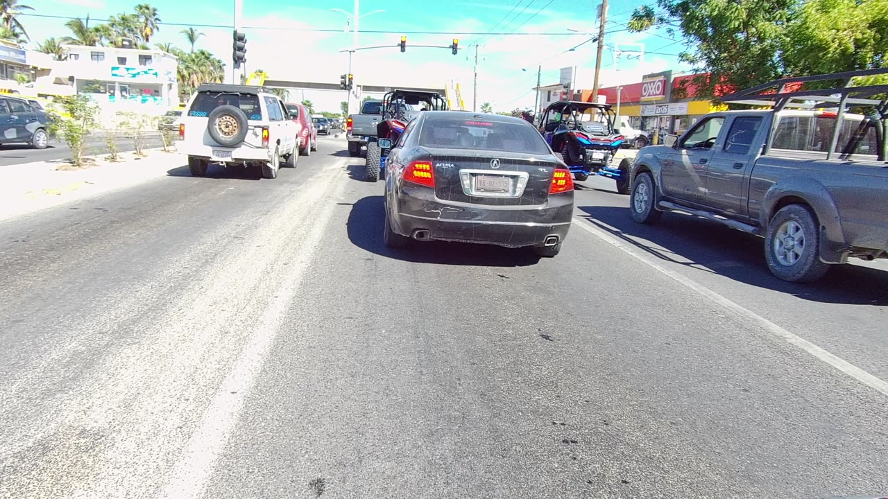 At 0:01, the stopped traffic line in Los Cabos remains nearly unchanged, with the black sedan centered, the white SUV slightly ahead, a gray pickup along the right lane, bright pavement glare, and no visible people.