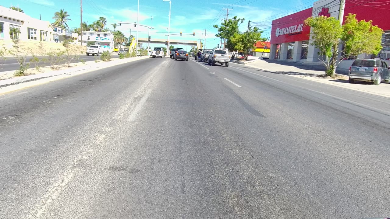 Opening frame of a Los Cabos street with asphalt lanes leading toward cars at traffic lights, parked vehicles, palm trees, low buildings, and a red storefront sign.
