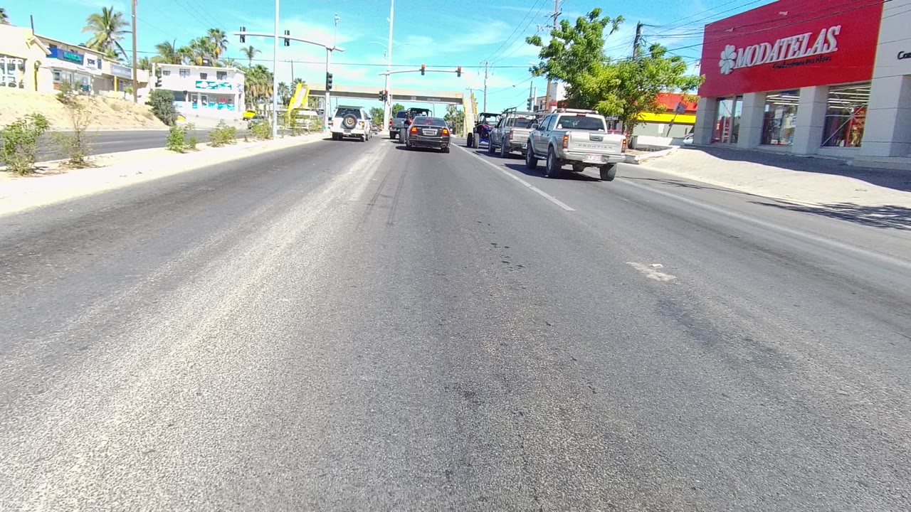 At 0:02, the Los Cabos roadway remains centered with dark pavement texture in front, cars clustered near traffic lights, and the red store facade more prominent at right.