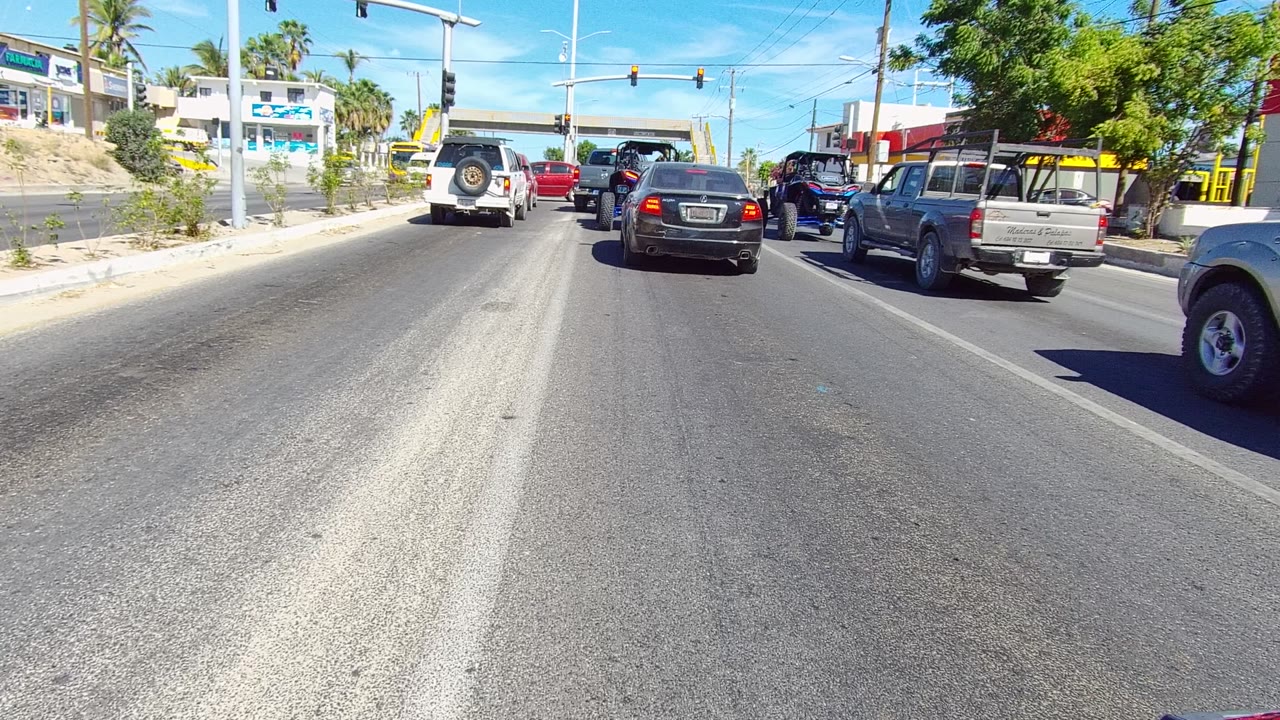 A white SUV, dark sedan, and pickup trucks sit near an overhead signal on a Los Cabos roadway, with roadside businesses, palms, and no visible people.