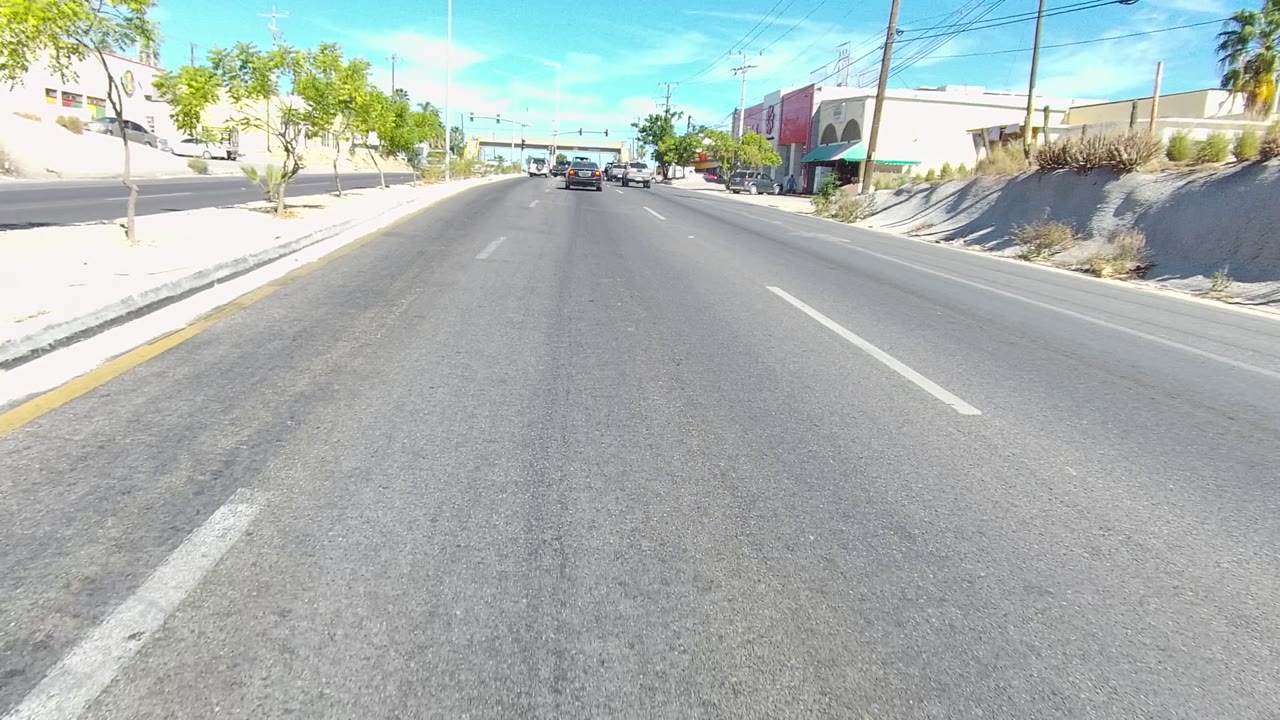 Forward road-level view on a sunny Los Cabos street with white lane markings, low buildings on both sides, parked vehicles, trees, power lines, and no visible people.