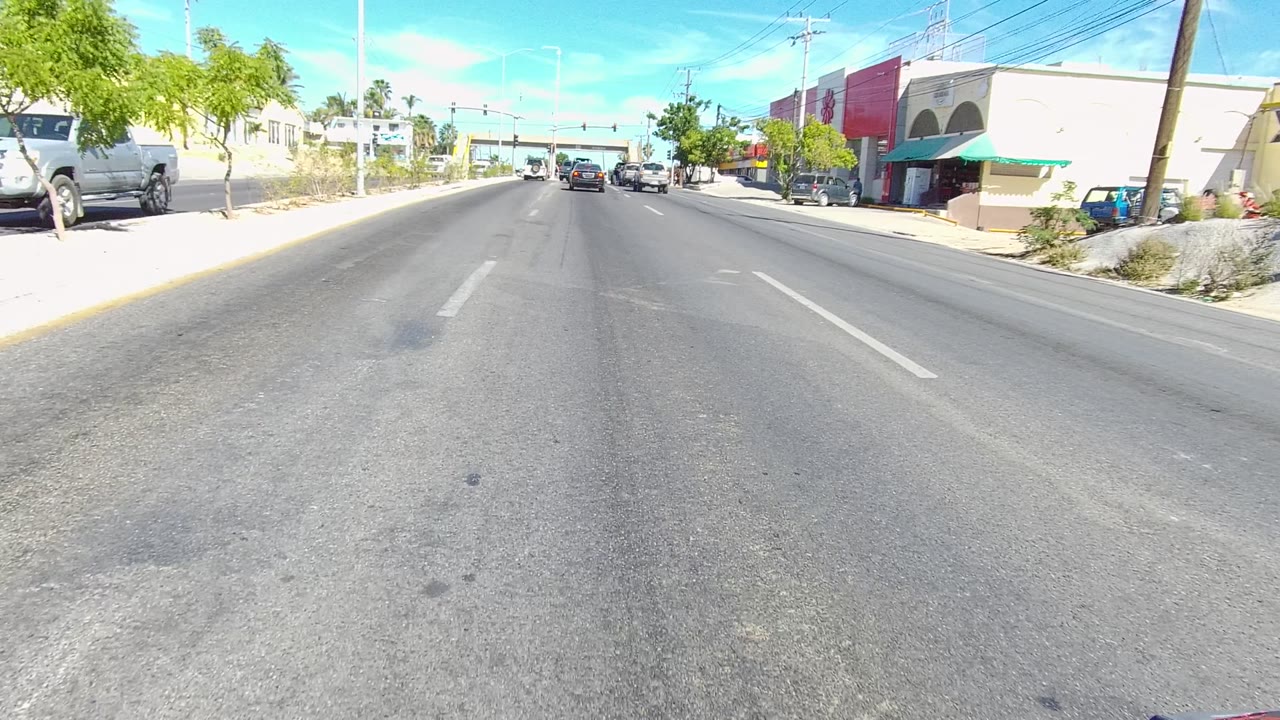 The Los Cabos roadway runs past a red and beige roadside building on the right, with parked vehicles, a sloped curbside area, power lines, bright sun, and no visible people.