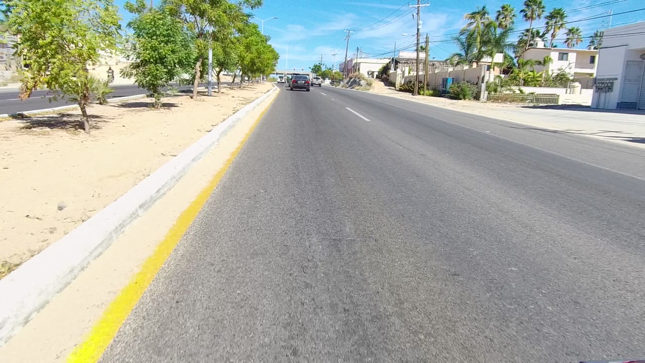 Opening frame of a Los Cabos road view with asphalt foreground, a yellow curb line at left, low buildings and trees on both sides, a car ahead, blue sky, and no visible people.