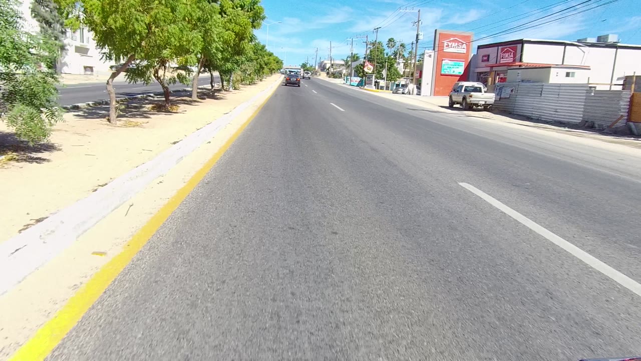 Opening frame of a Los Cabos city road with asphalt foreground, yellow curb line, trees, storefronts, vehicles in the distance, and no visible people.