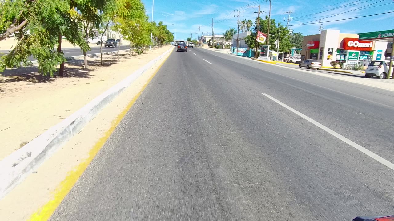 Los Cabos street frame with a red commercial building on the right, roadside trees, a sandy sidewalk, traffic ahead, overhead wires, and no visible people.