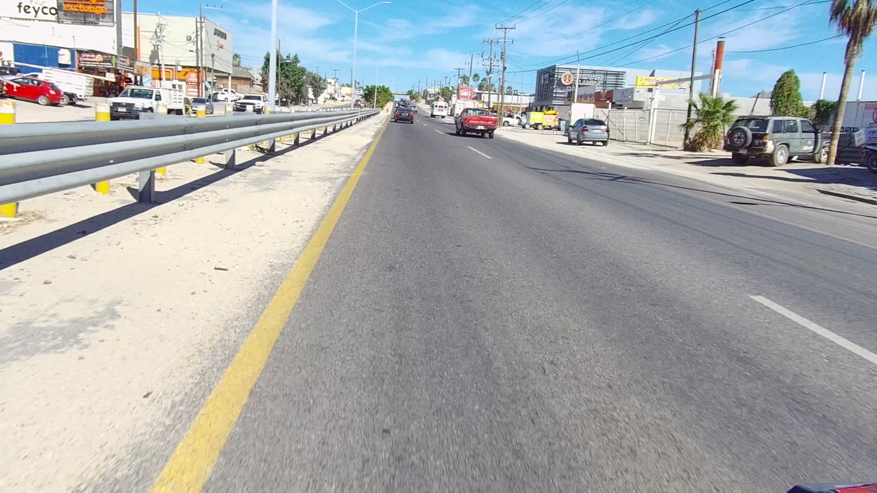Start of a Los Cabos road view with a metal guardrail on the left, cars ahead, roadside businesses, utility poles, and bright blue sky.