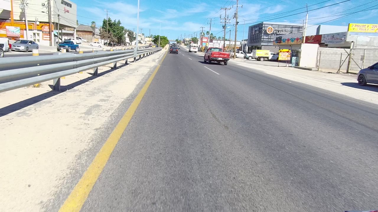 Forward road view with red pickup, guardrail, buildings, and overhead wires.