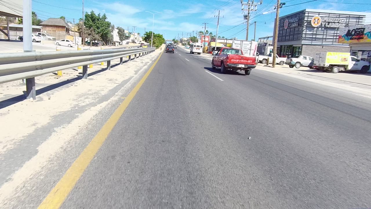 Mid-clip Los Cabos road view with cars moving ahead, a guardrail and sandy shoulder at left, storefront signage at right, and utility poles under clear sky.