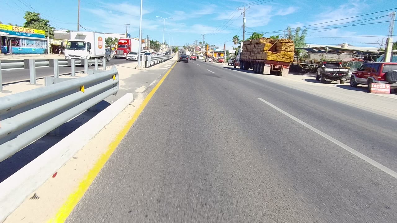 Opening road-level view in Los Cabos, Baja California Sur, with a metal guardrail on the left, yellow shoulder line, trucks and cars near roadside shops, and no clearly visible people.