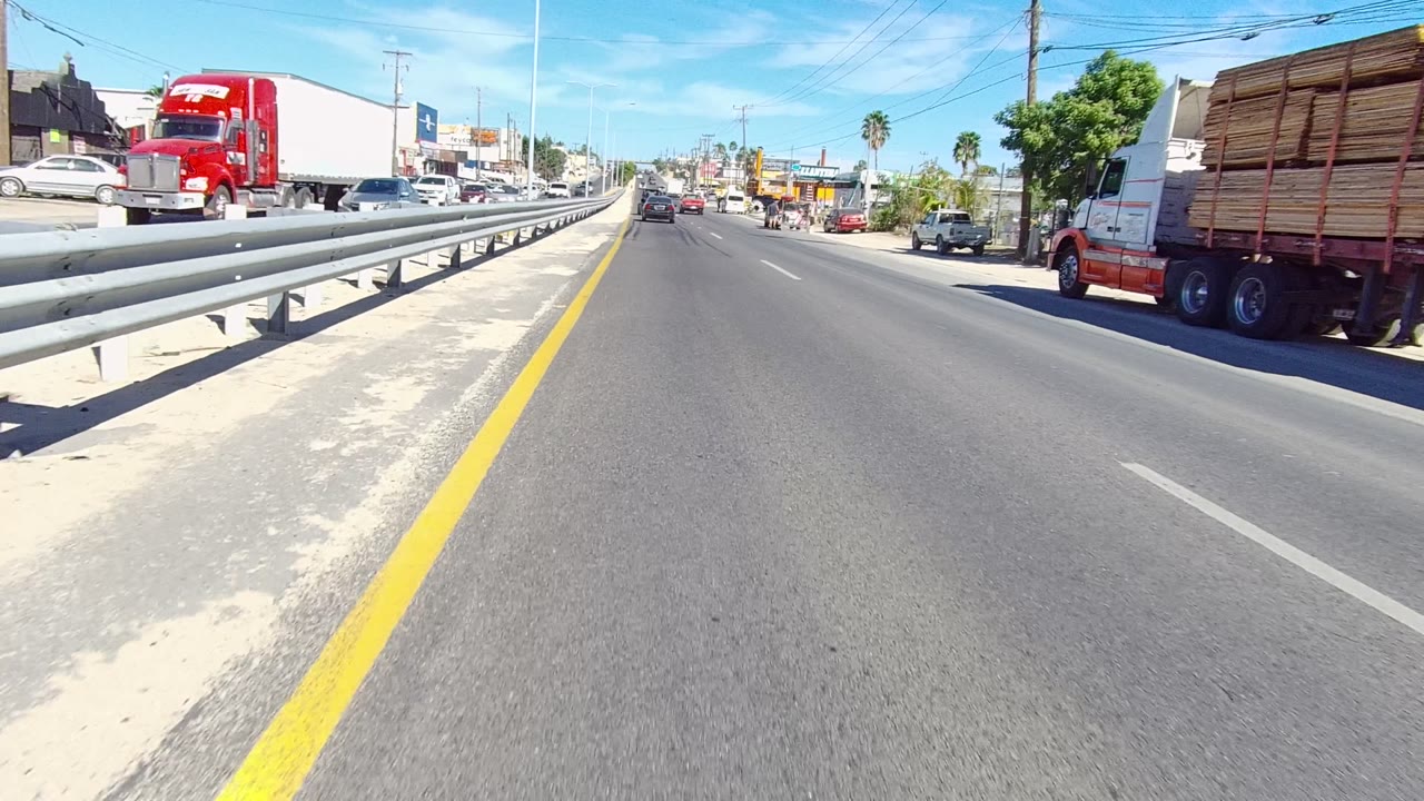 Forward road view with guardrail, red semi truck, storefronts, parked cars, and palm trees.