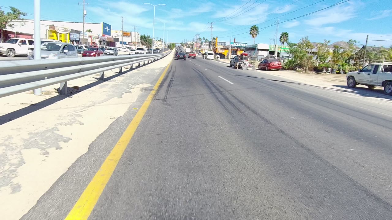 Mid-clip Los Cabos road frame showing the guardrail running forward, a truck loaded with stacked material on the roadside, cars in traffic, commercial buildings, palm trees, and no clearly visible people.