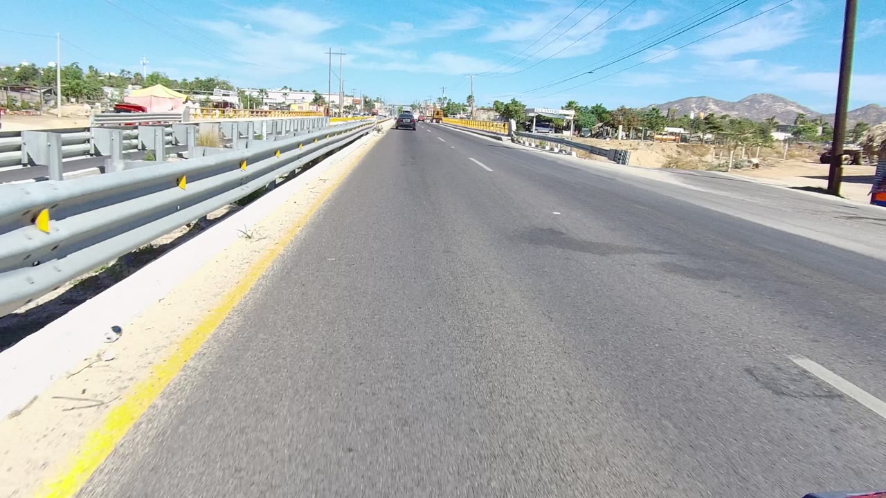 A road-level view on a Los Cabos bridge shows gray guardrails close to the lane, cars ahead, roadside buildings, mountains in the distance, and no visible people.