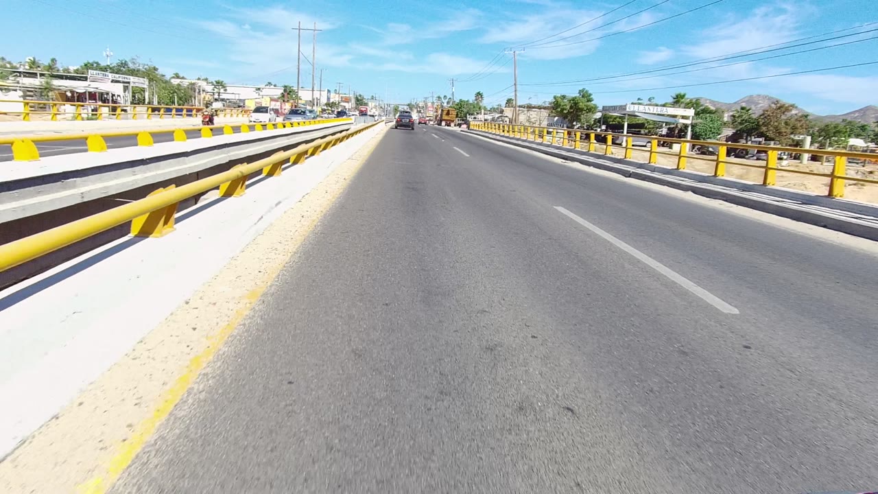 A forward road view in Los Cabos shows yellow bridge guardrails dominating the left side, traffic ahead, roadside structures, power lines, distant hills, and no visible people.