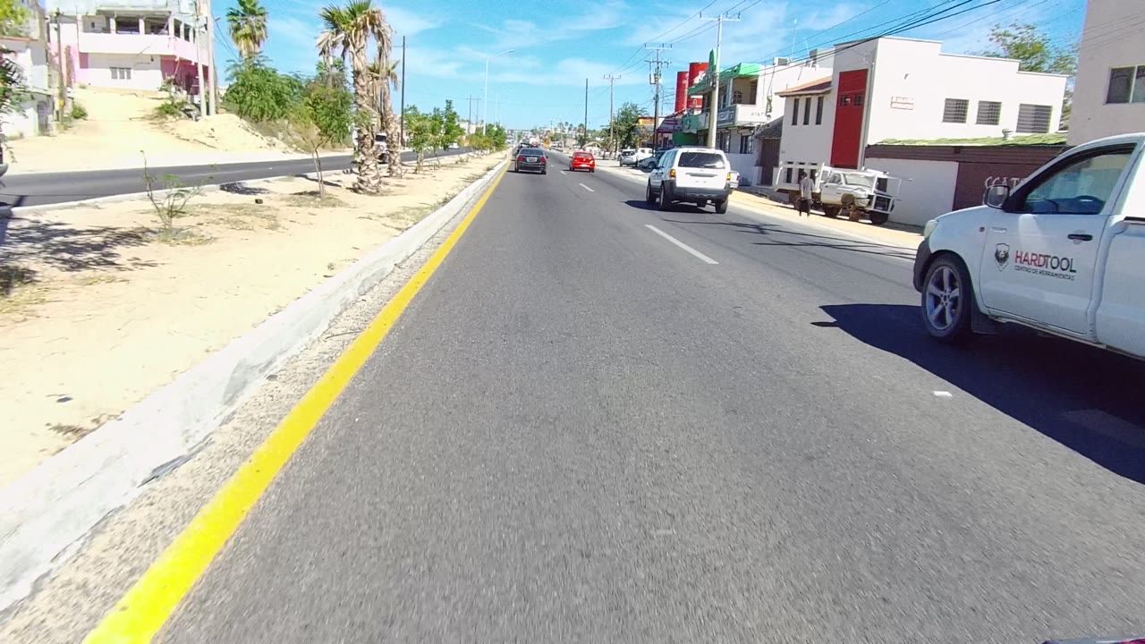 A Los Cabos road scene shows a forward view past a yellow curb line, sandy roadside, palms, storefronts, and moving cars, with no visible people.