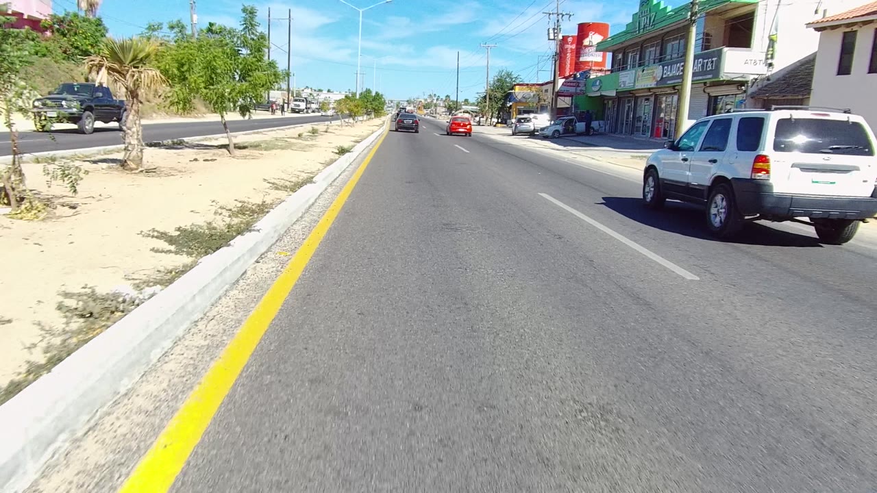 Urban roadway with a white SUV on the right, sandy median, palm trees, and storefronts ahead.