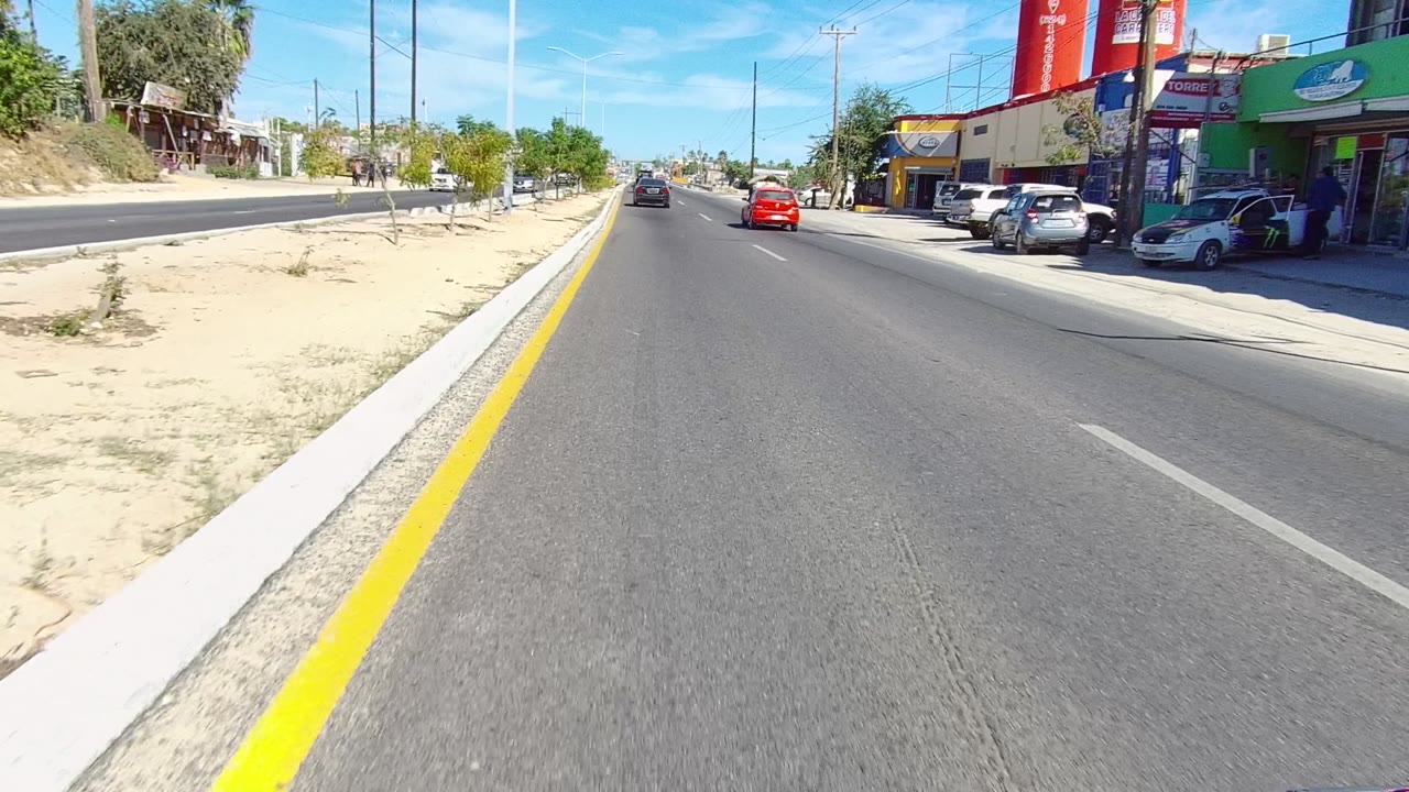 A Los Cabos street view looks straight down the roadway toward a red car, with storefronts and parked vehicles to the right and sandy divider planting to the left, with no visible people.